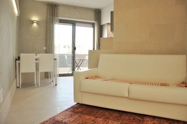 A modern living room with a white sofa, a dining table with chairs, and a balcony with a view.
