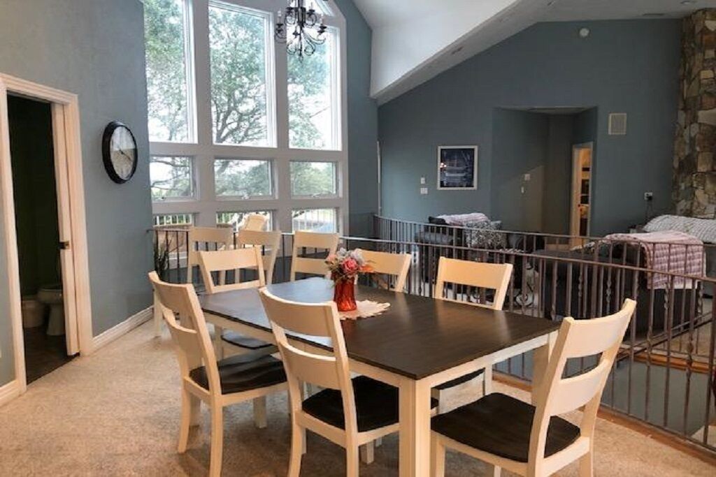 Un coin repas avec une table dressée pour six personnes, entourée de chaises, dans une pièce dotée de grandes fenêtres et d’un mur en pierre servant d’élément décoratif.