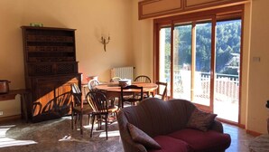 A dining area with a wooden table and chairs, a sofa, a bookshelf, and a view through large sliding glass doors.