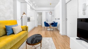 Una sala moderna con un sofá amarillo, una mesa de centro negra y un televisor. Hay una zona de comedor con una mesa blanca y sillas azules. La habitación tiene paredes blancas y suelo de madera.
