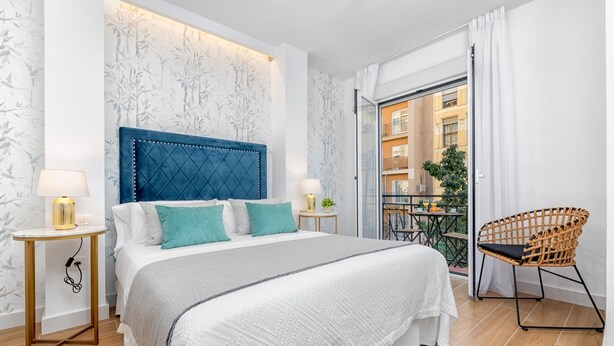 Un dormitorio moderno con una cama grande, cabecera azul, balcón con una silla de mimbre y vistas a edificios.