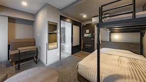 Quarto de hotel compacto com beliches, área de estar, chuveiro e uma pequena mesa de trabalho.