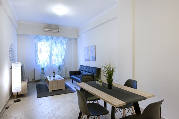 A modern living room with a sofa, a coffee table, and a dining table with chairs. There is a window with sheer curtains, a radiator, and a wall-mounted air conditioner. - Comfortable Apartment At The Foot of The Odeon of Herodes Atticus (Athens)