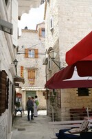 Uma rua estreita com edifícios de pedra, um toldo vermelho e uma placa indicando um restaurante.