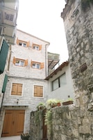 Uma rua estreita com edifícios de pedra, persianas de madeira e uma pequena área de jardim com vasos de plantas.