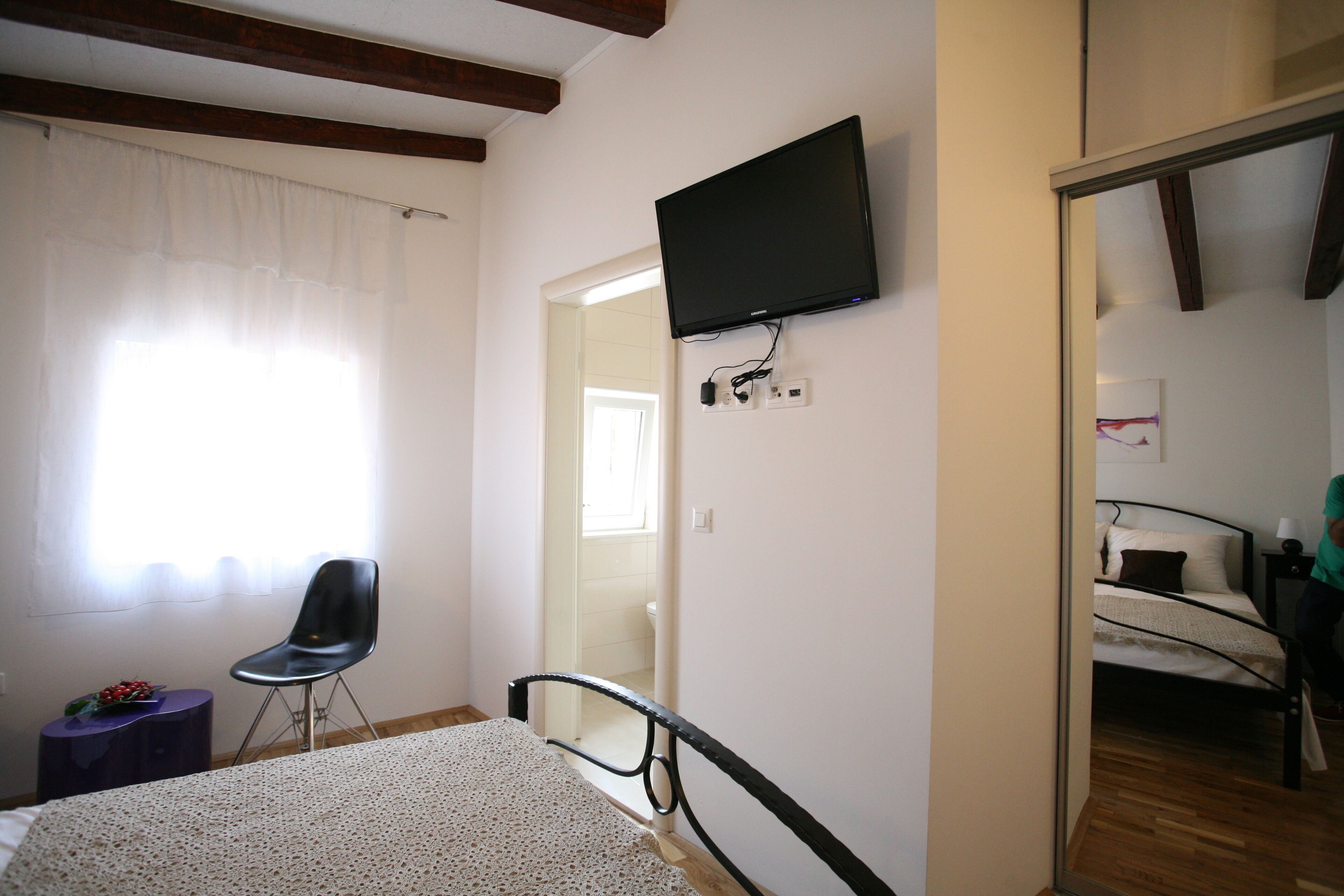 A bedroom with a bed, a chair, a television mounted on the wall, and a door leading to another room.