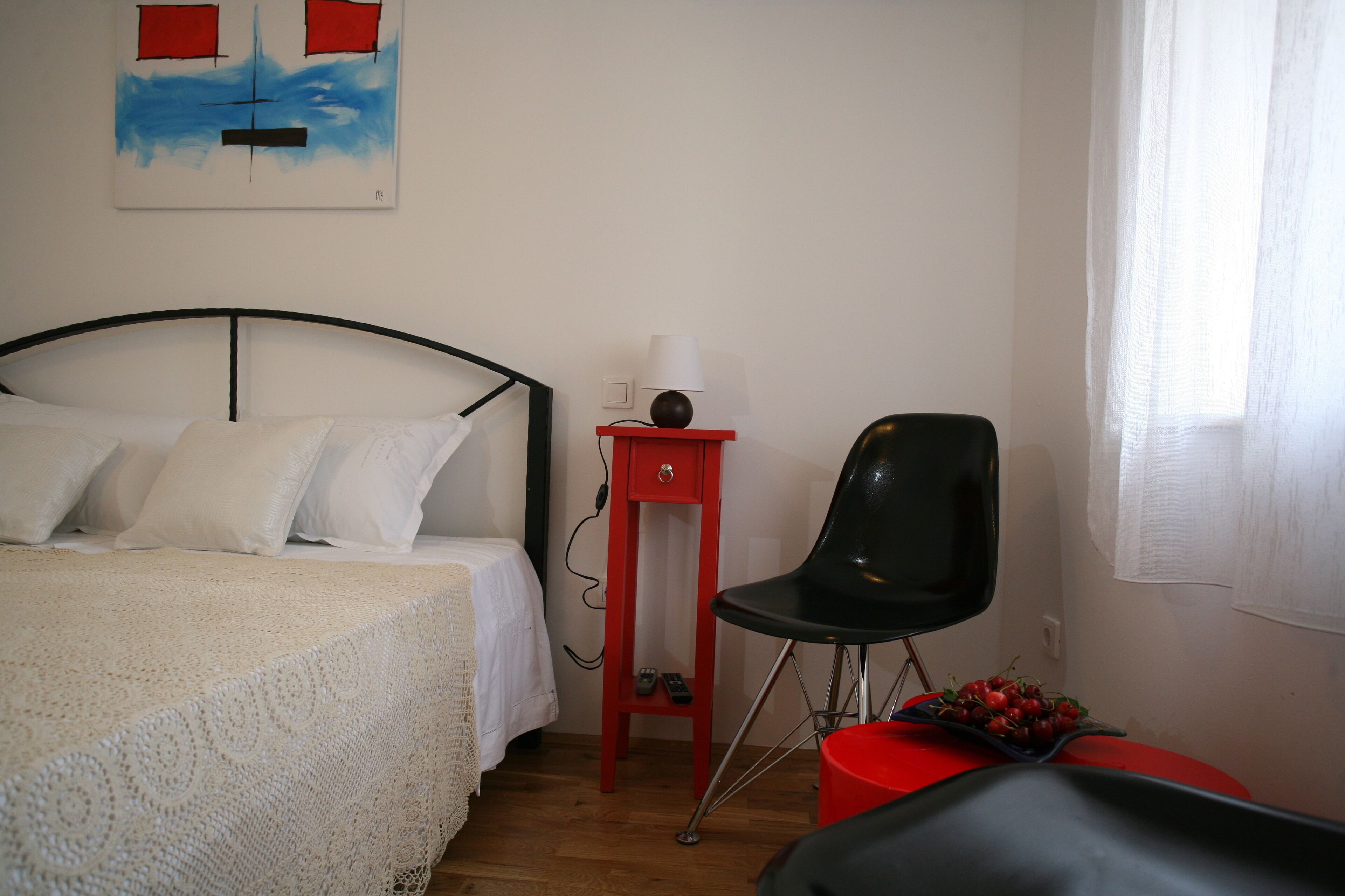 A bedroom with a bed, a red nightstand, a black chair, and a painting on the wall.
