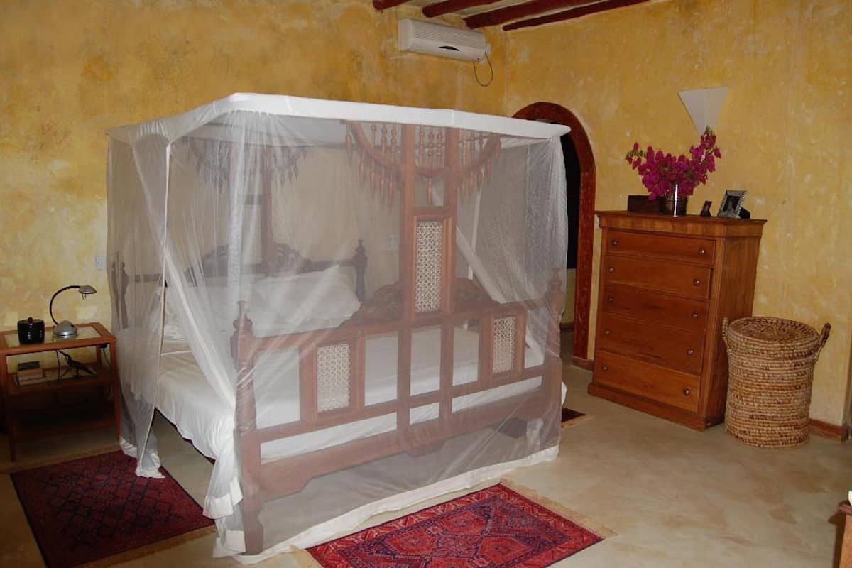 A room with a bed covered by a mosquito net, a wooden dresser, a lamp, and a wicker basket.