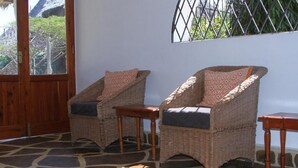 Two wicker chairs with cushions and a small table, a stone floor, and a window with a view of greenery. - Lighthomez Villa Resort (Mombasa)