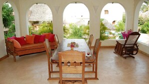 A covered dining area with a table and chairs, a sofa with cushions, and a view of a garden through arched openings. - Lighthomez Villa Resort (Mombasa)