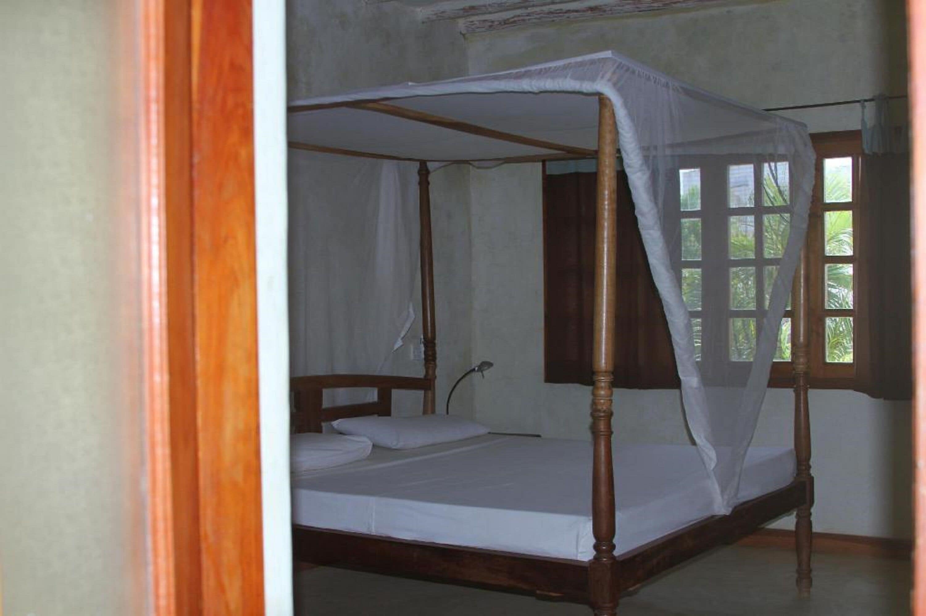 A four-poster bed with a canopy, a single bedside table, and a window with a view of greenery.