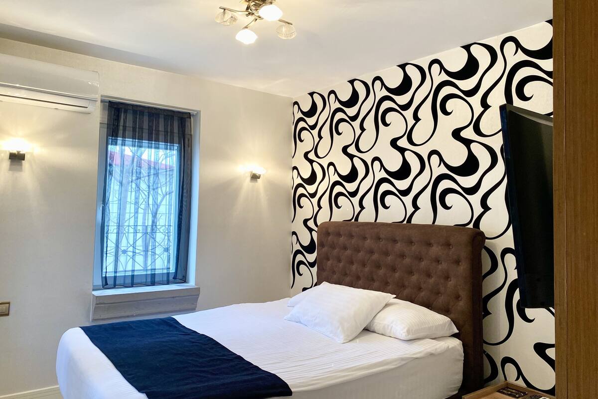 A bedroom with a bed, a window with curtains, a wall with a black and white pattern, and a ceiling light fixture.