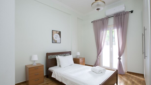 A neatly arranged bedroom with a wooden bed, a bedside table with a lamp, a small drawer unit, and a window with curtains. - Two Bedroom Apartment at Koukaki (Athens)