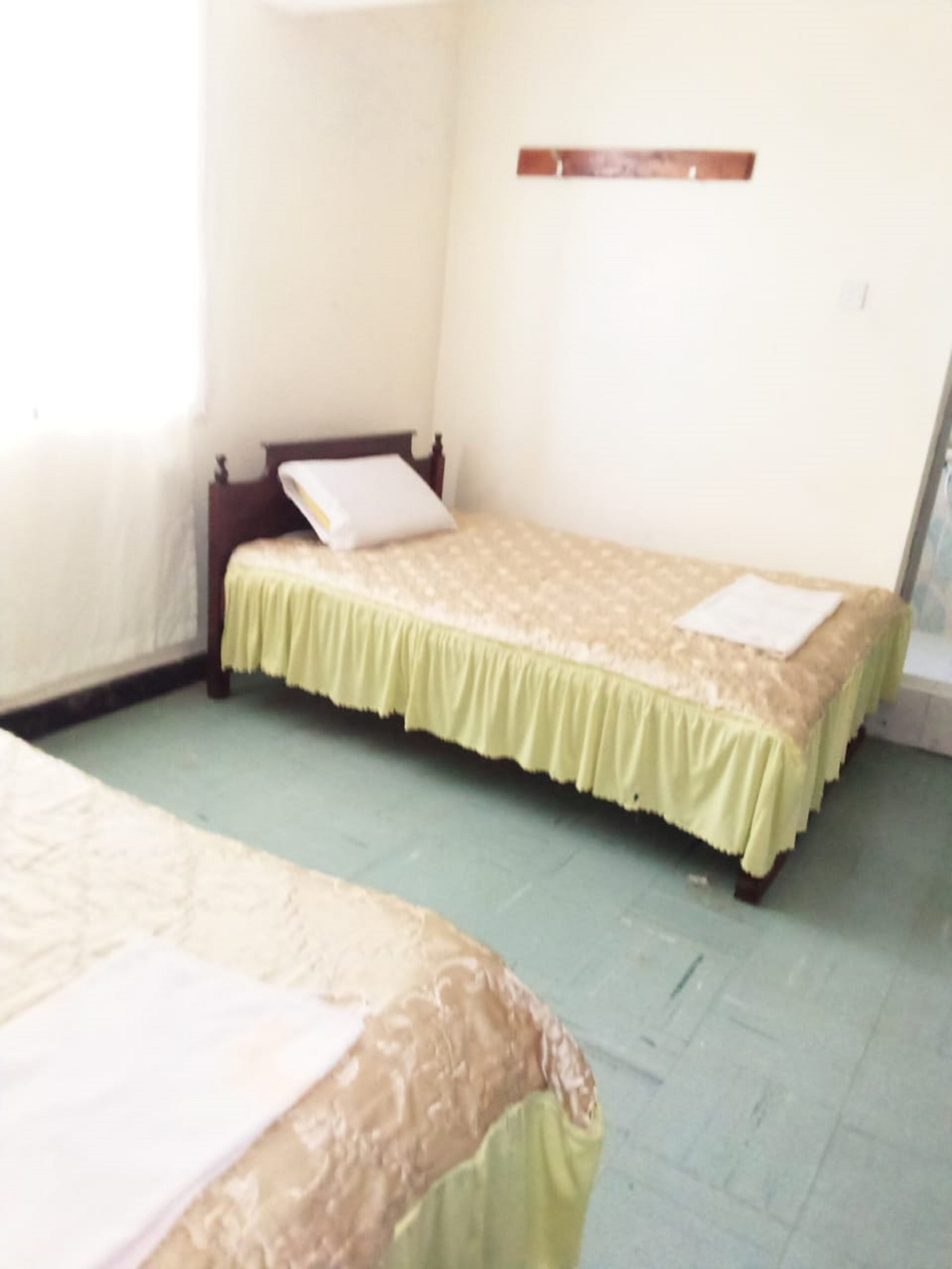 A room with two beds, each with a green bedspread and a white pillow. There is a wooden headboard on one bed and a wooden shelf on the wall. The room has a light green floor and white walls.