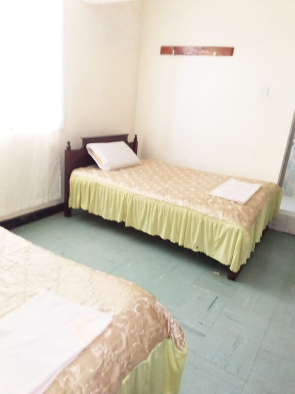 A room with two beds, each with a green bedspread and a white pillow. There is a wooden headboard on one bed and a wooden shelf on the wall. The room has a light green floor and white walls. - Manguo Lodge (Nyahururu)