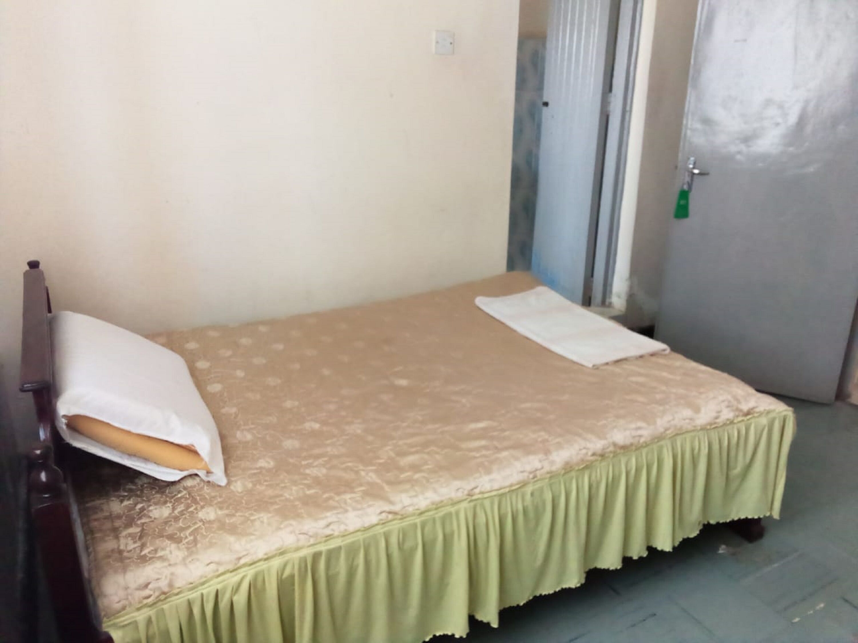 Une petite chambre avec un lit simple, une couvre-lit verte et un oreiller blanc. On peut voir une tête de lit en bois sur le côté gauche.