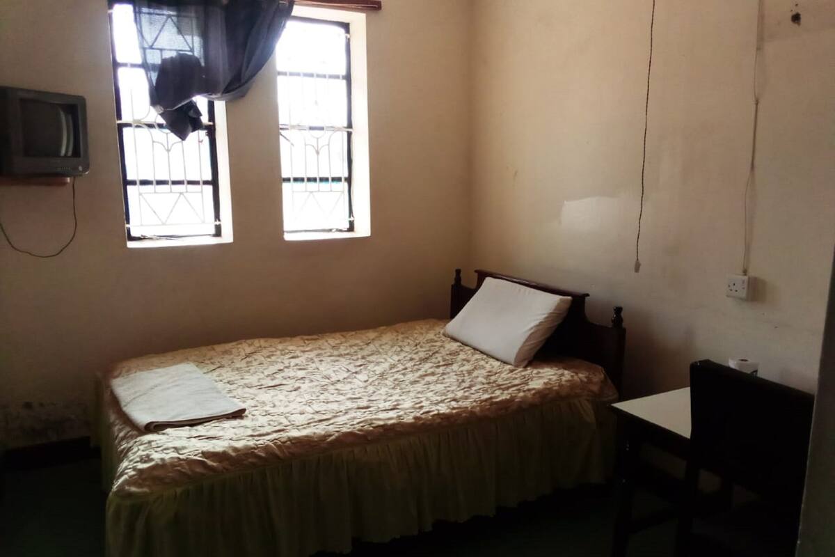 Una habitación pequeña y con poca luz, con una cama individual, una silla, un televisor y una ventana con barrotes.