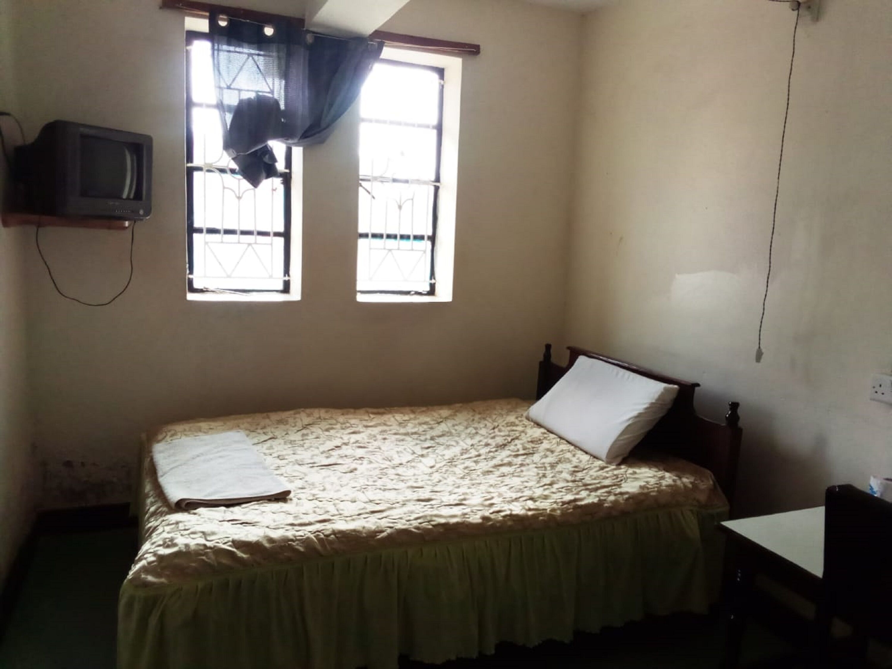 Una habitación pequeña y tenuemente iluminada, con una cama individual, un televisor montado en la pared y una ventana con barrotes.