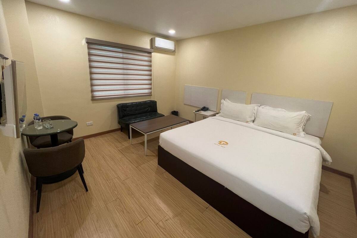 Ein Hotelzimmer mit einem Bett, einem Schreibtisch, einem Sessel, einem kleinen Tisch, einer schwarzen Ledersofa, einem Fenster mit Jalousien und einer Klimaanlage.
