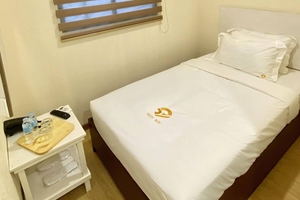 Ein Einzelbett mit weißem Kopfteil, einem Kissen, einem Nachttisch mit Fernbedienung, einer Wasserflasche und einem Glas, sowie ein Fenster mit Rollläden.
