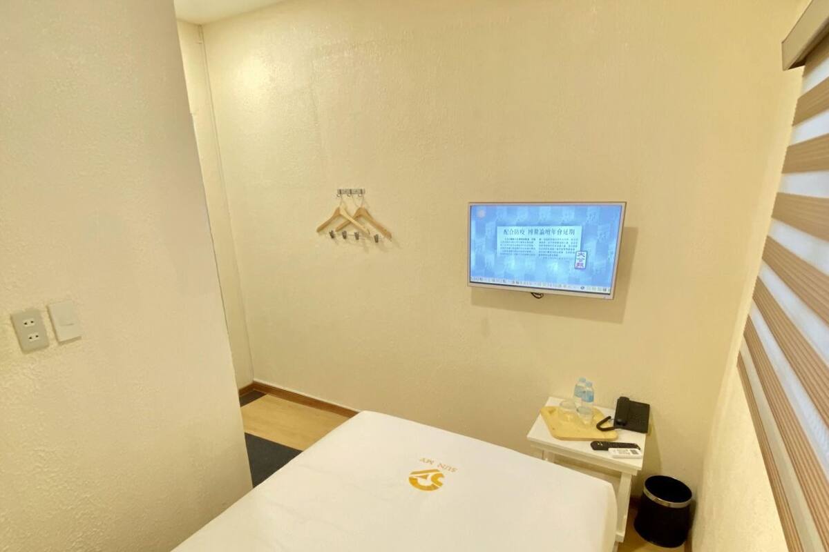 Ein Hotelzimmer mit einem Bett, einem an der Wand befestigten Fernseher, einem kleinen Tisch mit Telefon und Wasserflasche sowie einer Wand-Klimaanlage.