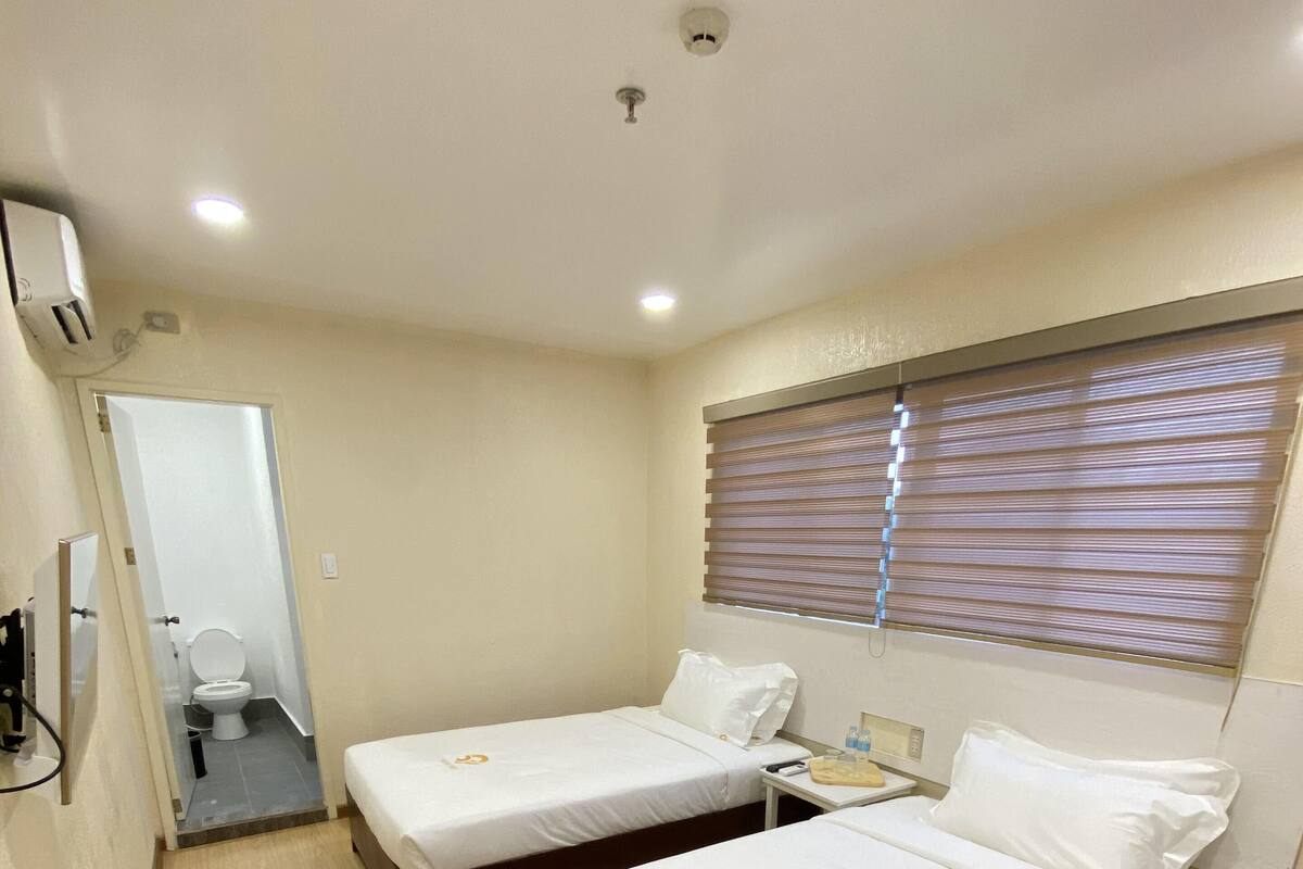Ein Hotelzimmer mit zwei Einzelbetten, einem kleinen Nachttisch mit Lampe, einem Fenster mit Jalousien, einer sichtbaren Toilette hinter einer offenen Tür und einer Deckenleuchte.