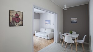 A modern living room with a white sofa, a dining table with chairs, and a small plant. There is a painting of Paris on the wall. - Zan Moreas A Simple & Minimal Apartment (Athens)