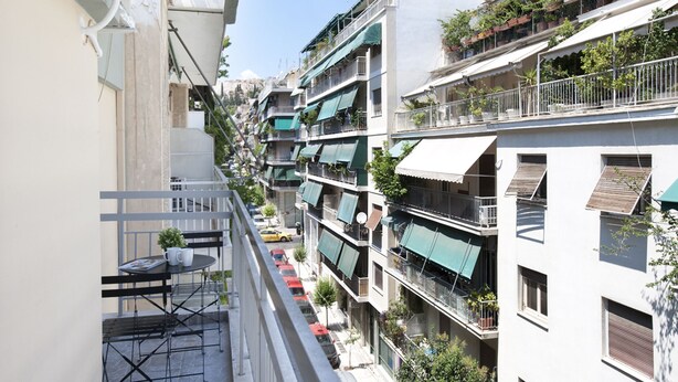A narrow urban street with multi-story buildings, balconies with plants, and a small outdoor seating area with a table and chairs. - Zan Moreas A Simple & Minimal Apartment (Athens)