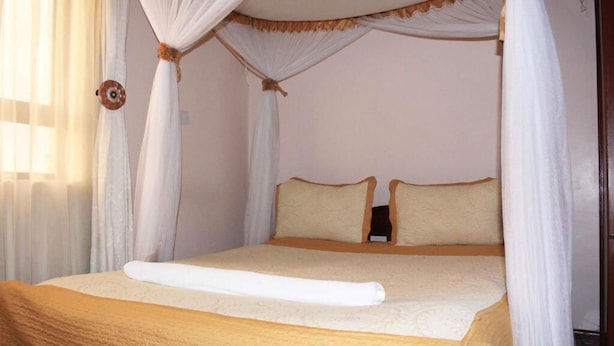 Un lit à baldaquin à quatre poteaux, avec une couvre-lit et des oreillers de couleur beige, une serviette blanche sur le lit et des rideaux transparents aux fenêtres.