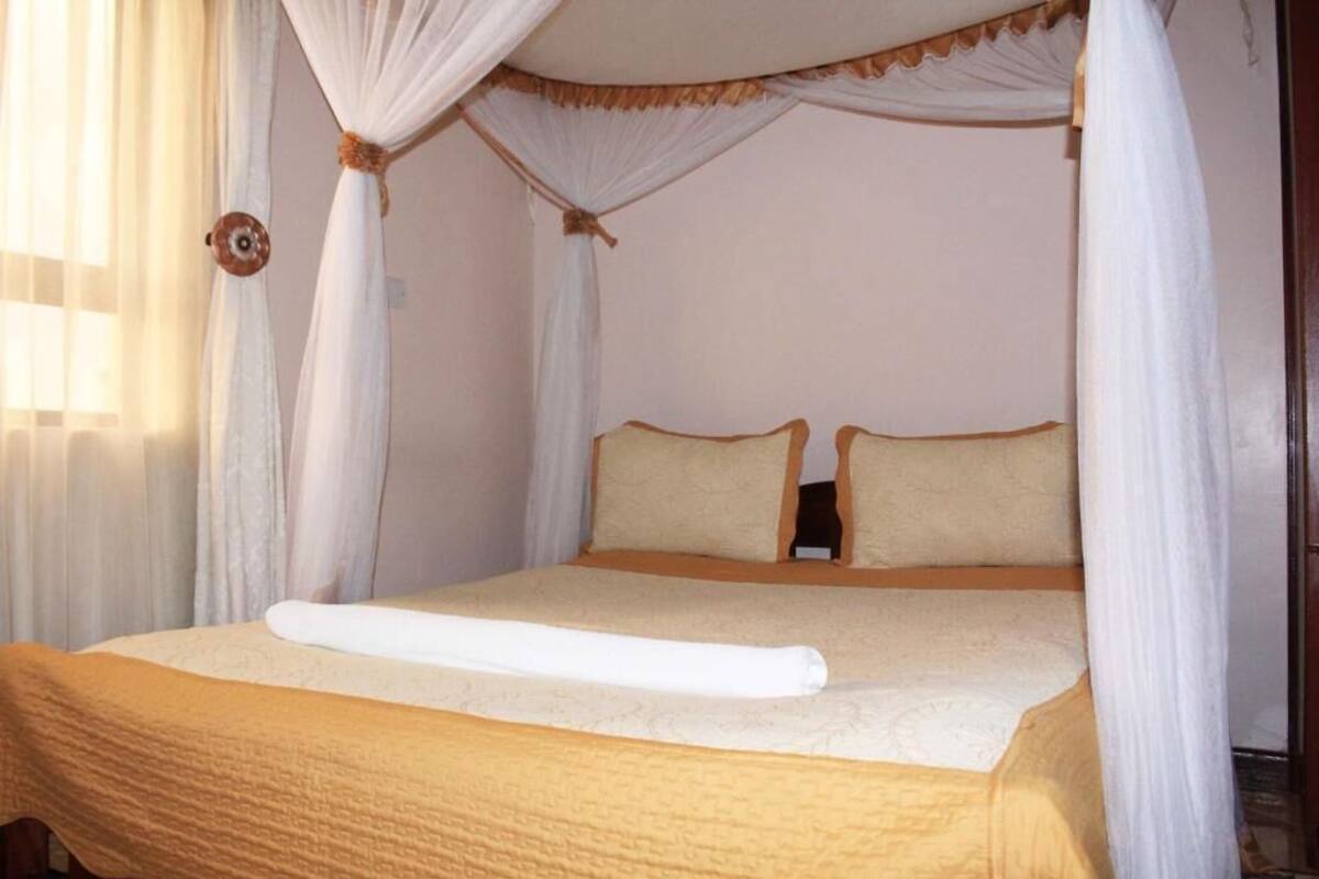 A four-poster canopy bed with a beige bedspread and pillows, a white towel on the bed, and sheer curtains on the windows.