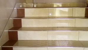 Un escalier dont les marches alternent entre des carreaux beiges et bruns, menant à un palier doté d’une rambarde en verre.