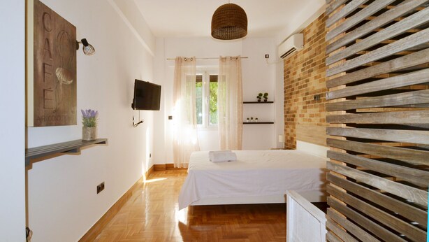 A bedroom with a bed, a television, a wooden slatted wall, a ceiling light, and a window with curtains.