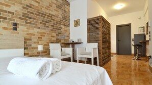 A hotel room with a brick wall, a bed with a rolled white towel, two white chairs, a small table, and a door.