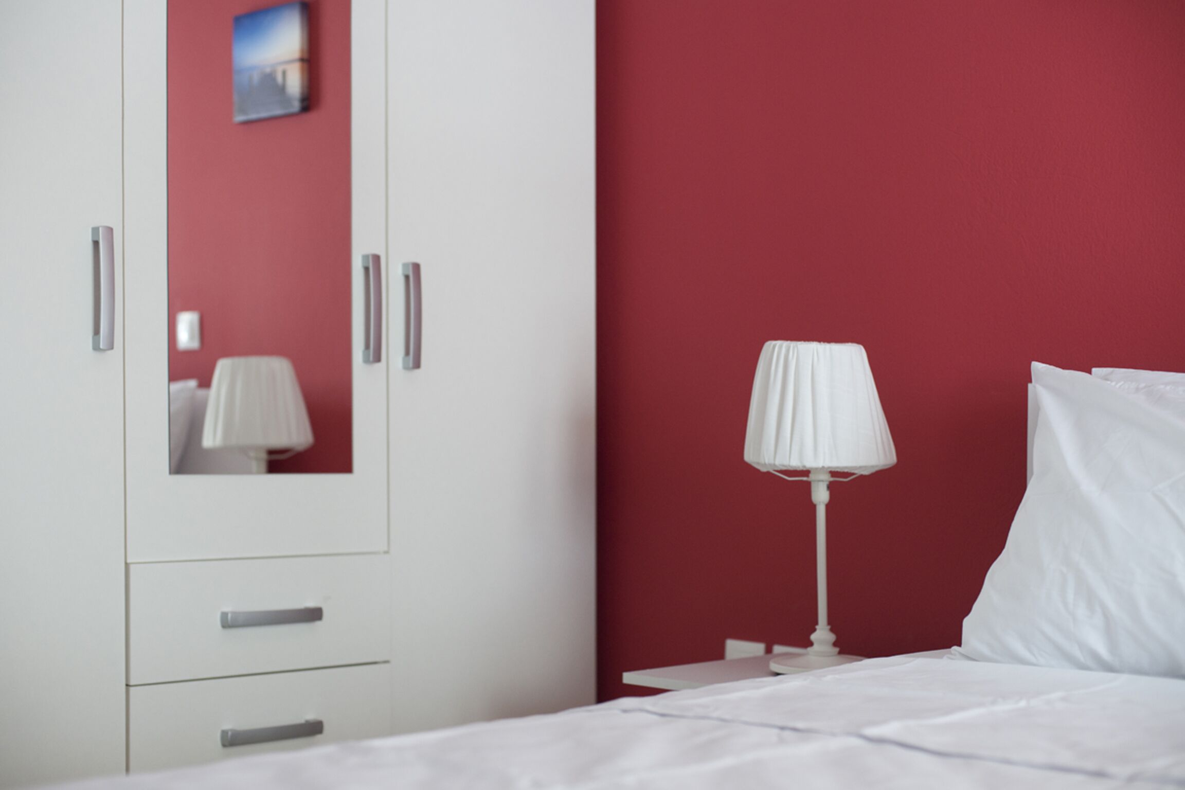 A hotel room with a bed, white curtains, a nightstand with a lamp, a wardrobe, and a red accent wall.