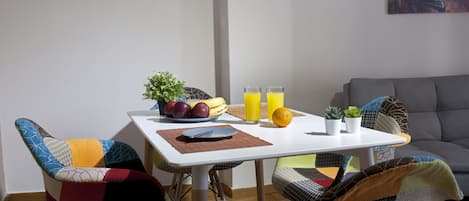 Un coin repas avec une table blanche, des chaises colorées et un saladier rempli de fruits.