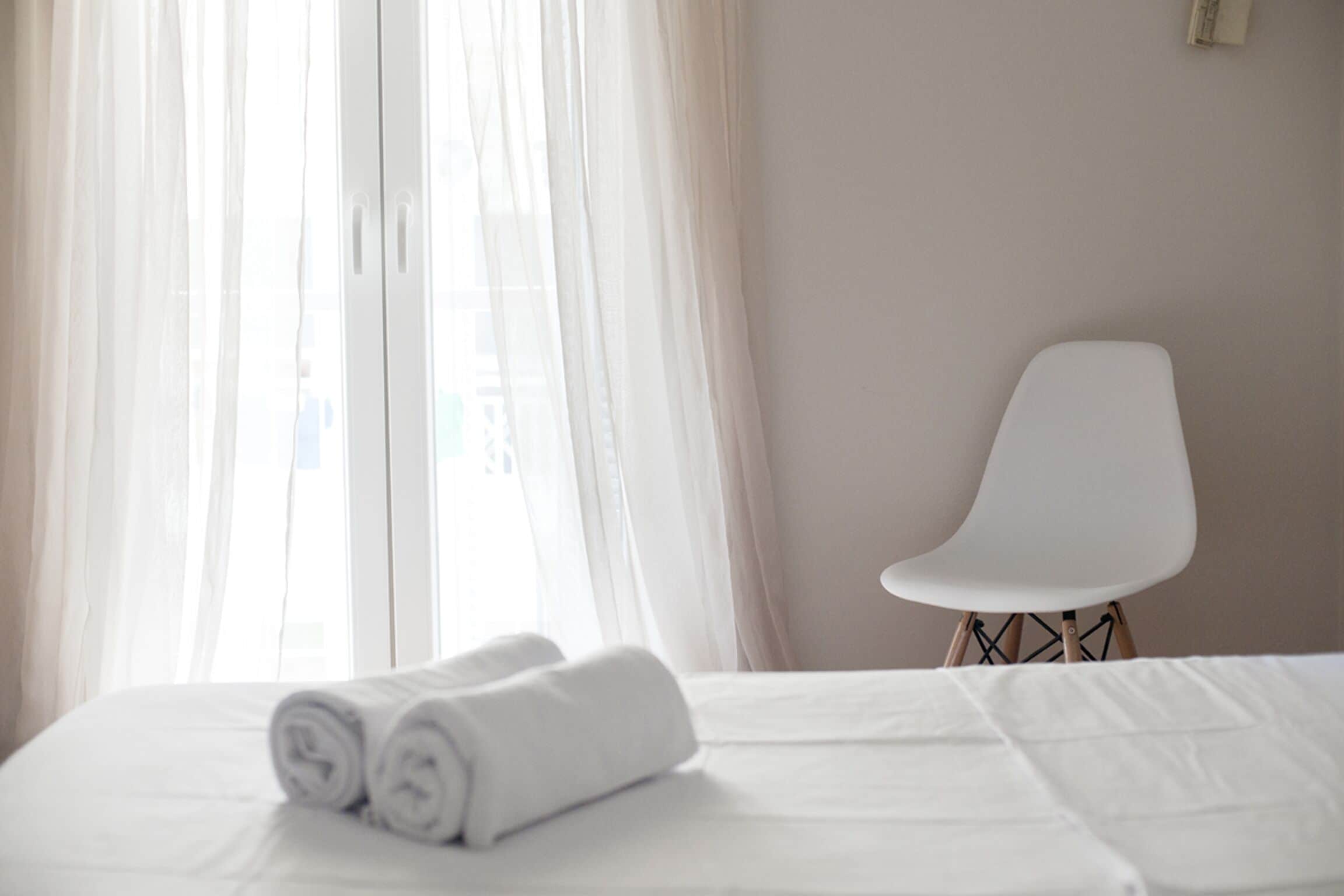 A hotel room with a bed, a rolled white towel, a white chair, and sheer curtains.