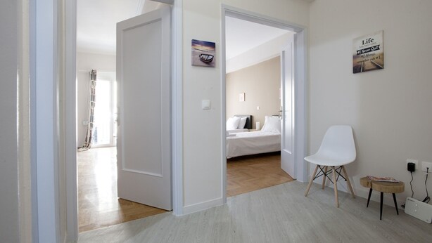 Ein modernes Zimmer mit einem Bett, einem weißen Stuhl und einem kleinen Tisch. Eine Tür führt in einen anderen Raum, und an der Wand befindet sich ein Bild sowie ein Schild.