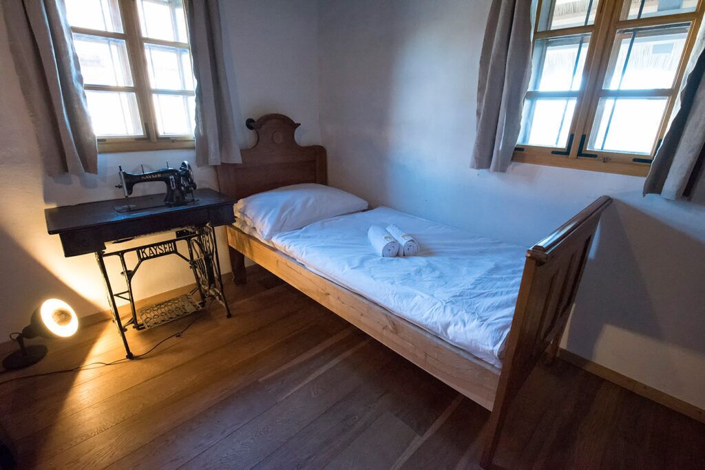 Uma cama de solteiro com cabeceira de madeira, uma máquina de costura sobre uma mesa e uma janela com cortinas.