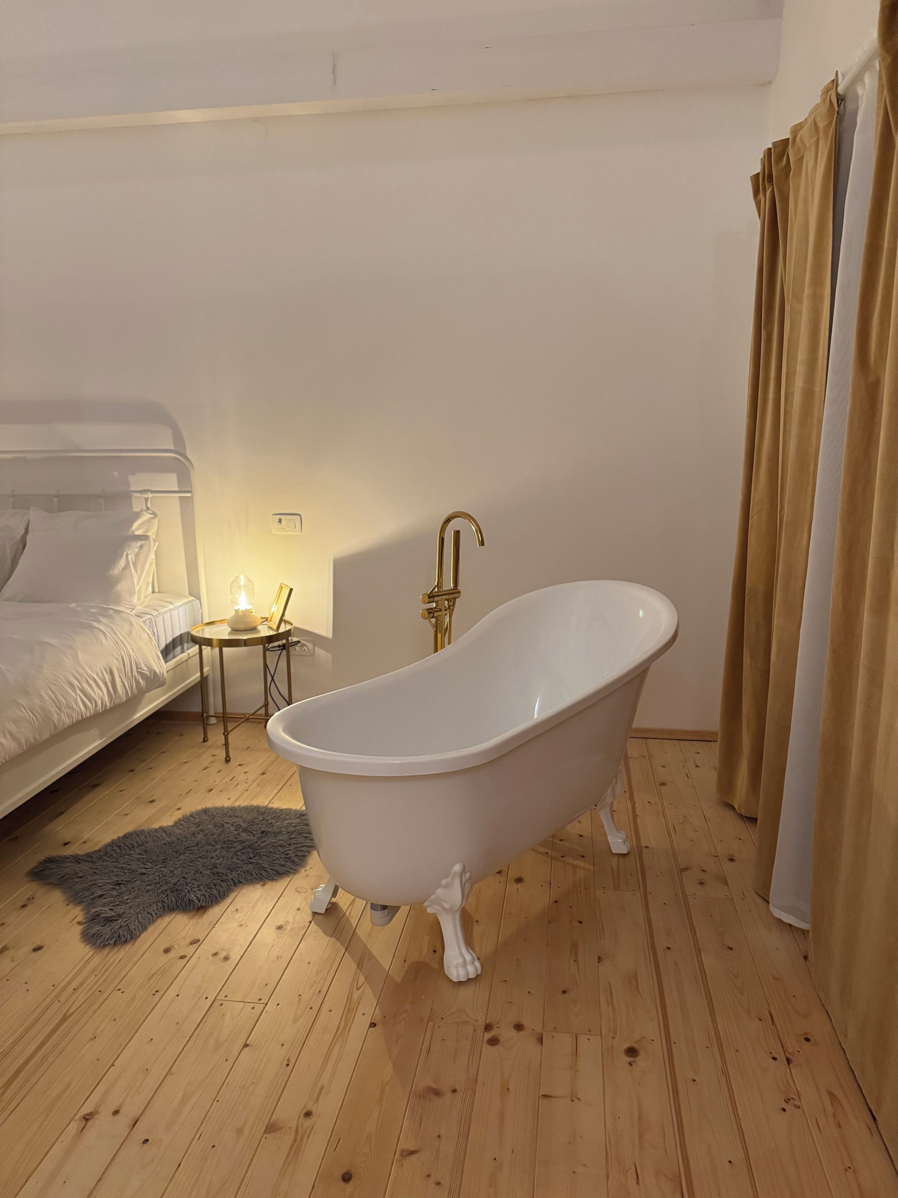 A room with a white bathtub, a bed with white bedding, a small bedside table with a lamp, and a grey rug on the wooden floor.