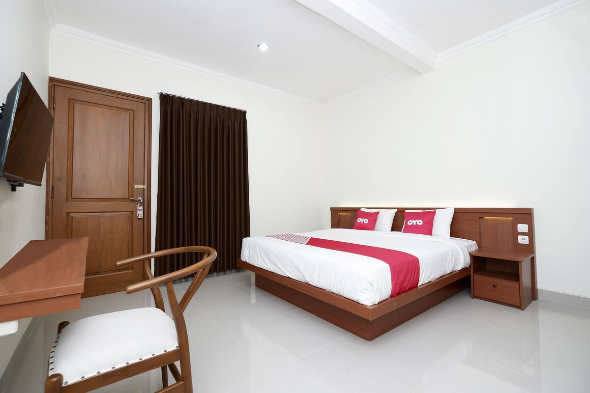 A hotel room with a bed, wooden nightstand, chair, and a desk with a flat-screen TV.