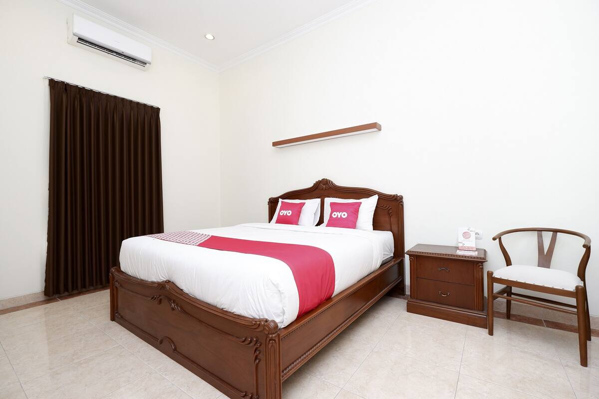 A neatly made bed with a wooden headboard, a bedside table, a chair, and a wall-mounted shelf.