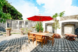 An outdoor patio with a wooden table and chairs, a red umbrella, and stone walls. - OYO 2327 Pagsis Village (Klaten)