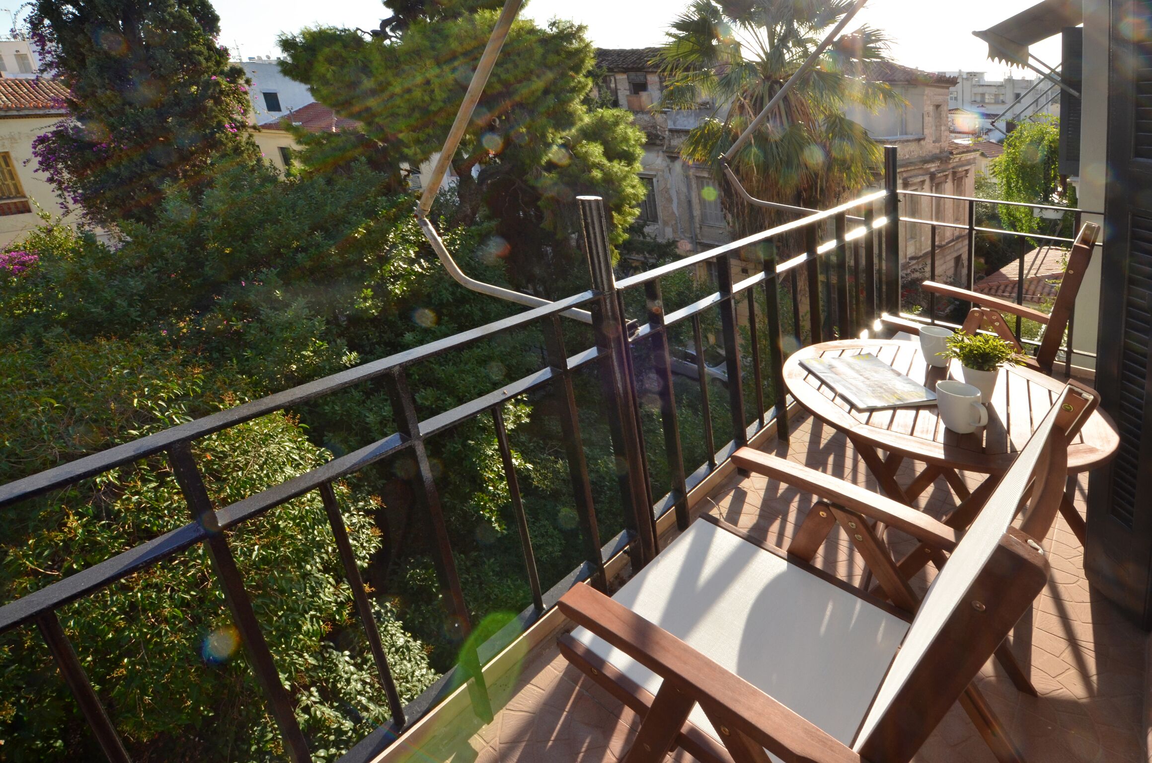En balkon med et træbord og stole, en plantekasse og udsigt til omkringliggende bygninger og træer.