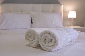 Una cama bien tendida con sábanas blancas y toallas enrolladas, un cabecero y una lámpara de noche.