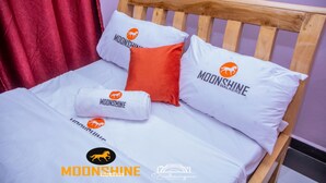 A bed with white bedding and pillows featuring the 'Moonshine Walkers' logo, a wooden headboard, and a red curtain. - Moonshine Walkers (Eldoret)