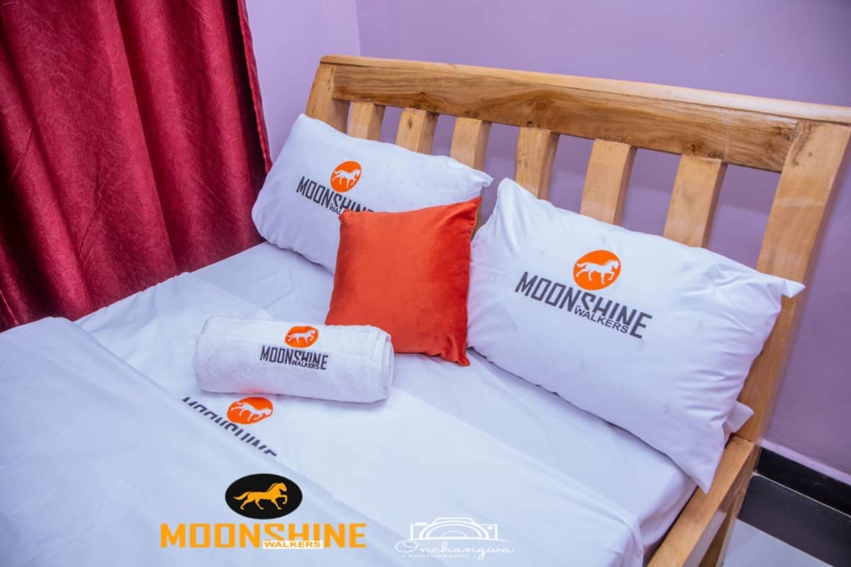 Ein Bett mit weißer Bettwäsche und Kissen mit dem 'Moonshine Walkers'-Logo, ein hölzernes Kopfstück und ein roter Vorhang.