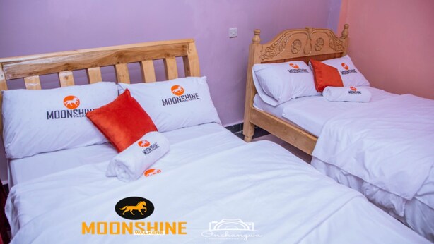 Two twin beds with wooden headboards, white bedding, and orange pillows with the Moonshine logo. - Moonshine Walkers (Eldoret)