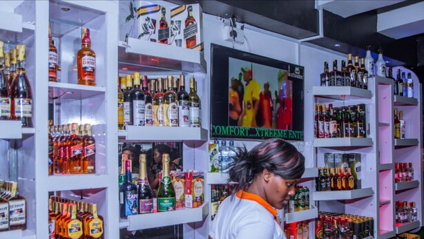A liquor store with shelves stocked with various bottles and a television screen displaying an advertisement. - Moonshine Walkers (Eldoret)