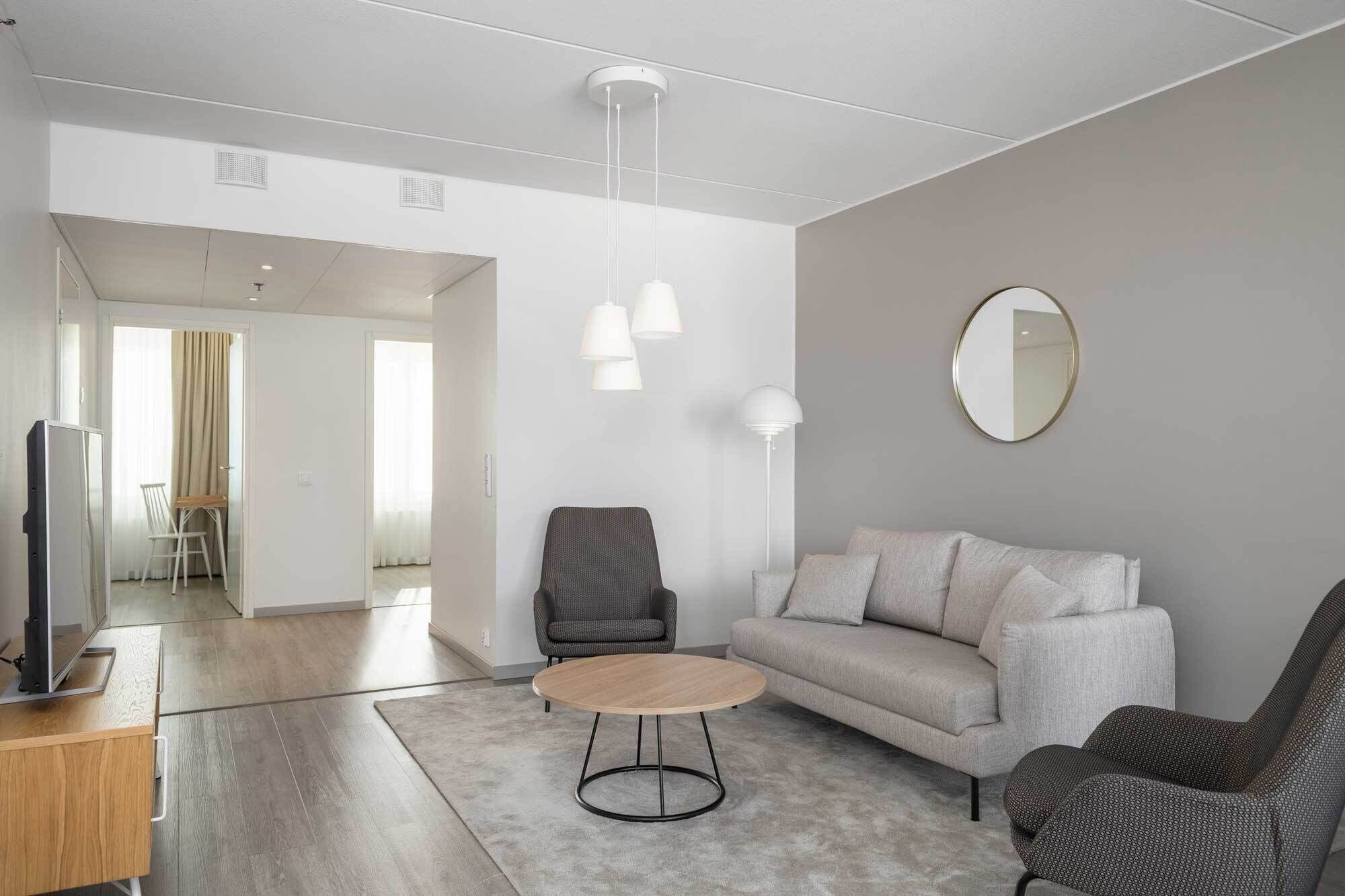 A modern living room with a sofa, two armchairs, a coffee table, and a TV. There is a mirror on the wall and a ceiling light fixture.