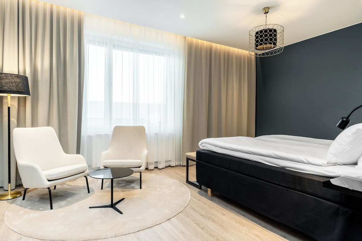 A modern hotel room with a bed, two armchairs, a small table, and a floor lamp.